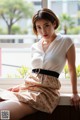 A woman sitting on a window sill in a white shirt and skirt.