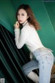 A woman in a white sweater and jeans leaning against a green wall.